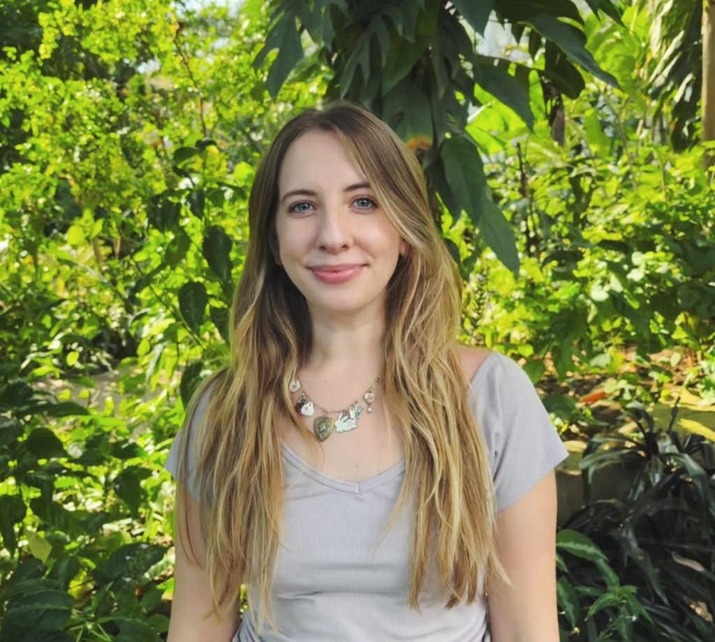 A woman with long, blonde wavy hair stands in front of green foliage, wearing a purple top and a charm necklace, smiling at the camera.